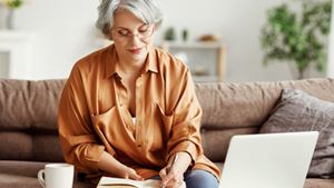 A retiree sitting on a sofa taking notes whilst working from their laptop