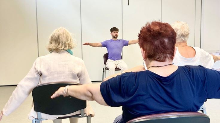 Personal trainer, Ben Broadhead, leading an older adult exercise workshop. Ben sits on a chair with his arms outstretched and the other people in the room copy his movements
