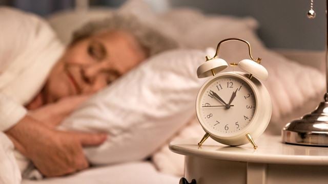 An older woman peacefully asleep in bed with an alarm clock in the foreground highlighting sleep needs for older adults.