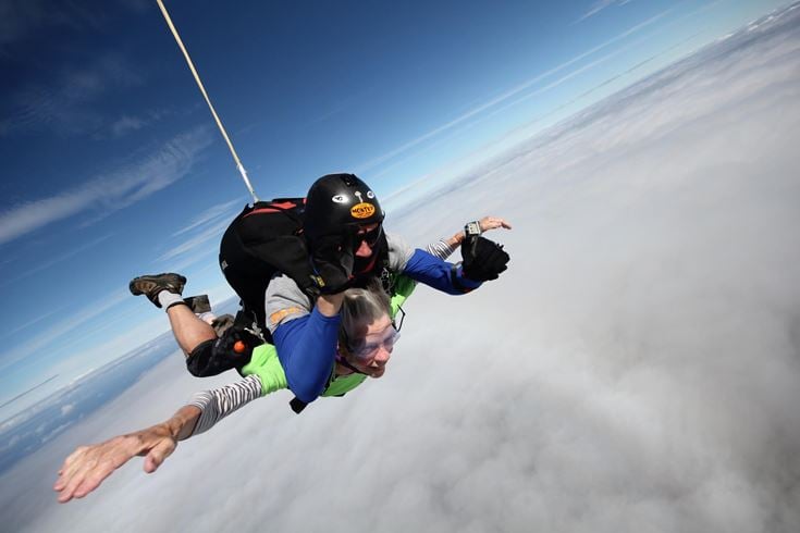 An 80-year-old senior skydiving, guided by a professional with a parachute attached to their back. They glide through the air above the clouds, arms outstretched with the wind in their faces