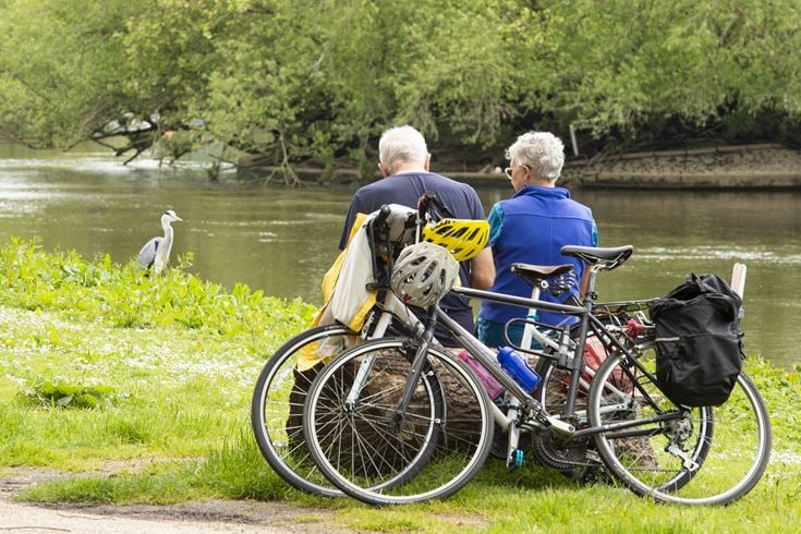 Two retired people taking a break from cycling by resting on a log by the river.