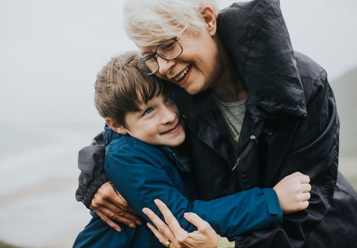 A grandmother and grandson smiling and hugging in a coastal location on a grey, wet looking day.