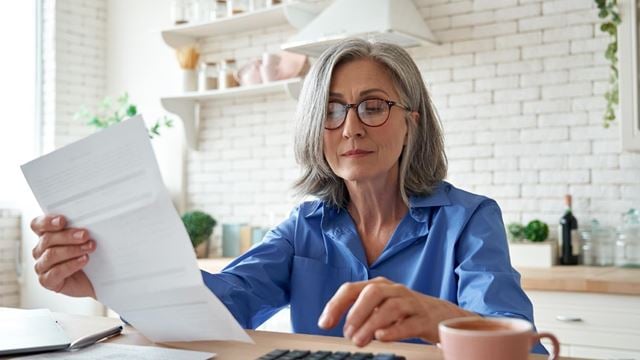 Senior woman reviewing finances with a calculator and paperwork at her laptop considering if pensioners pay tax