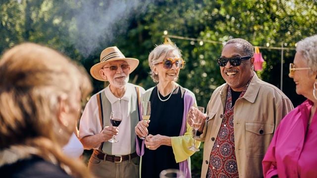 A group of older people enjoying a garden party, with various drinks in hand and smoke from a barbecue pluming overhead