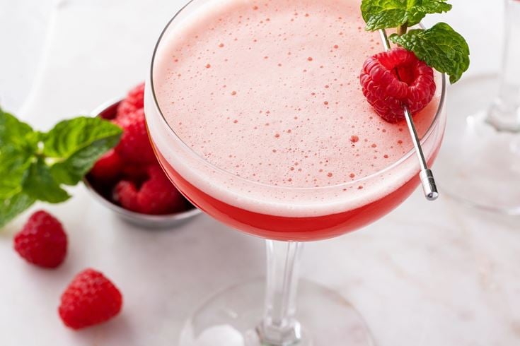 A glass of Rudolphs Tini cocktail on a marble worktop next to a bowl of raspberries.