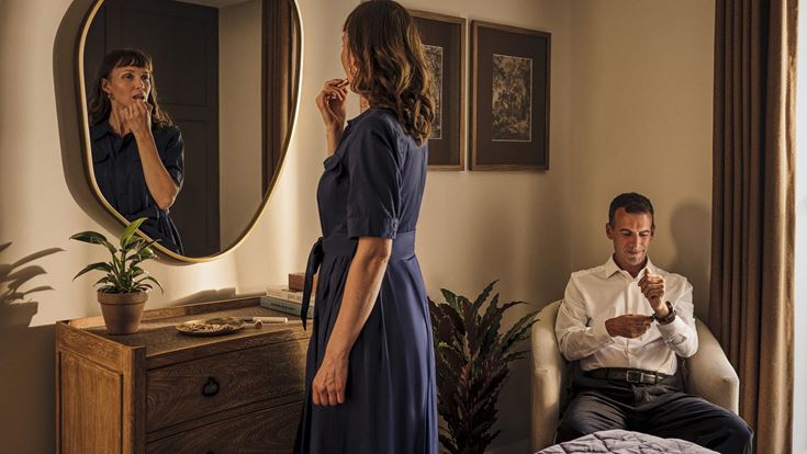 a woman standing in front of a mirror and a man sitting on a bed