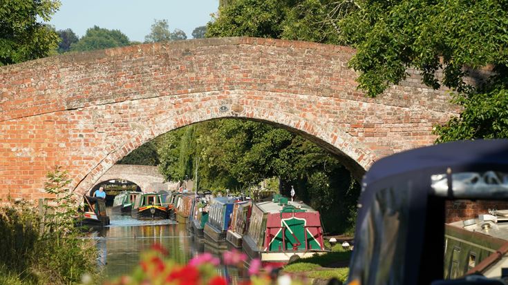 Colourful flowers in front of a canal lined with boats near a bridge.