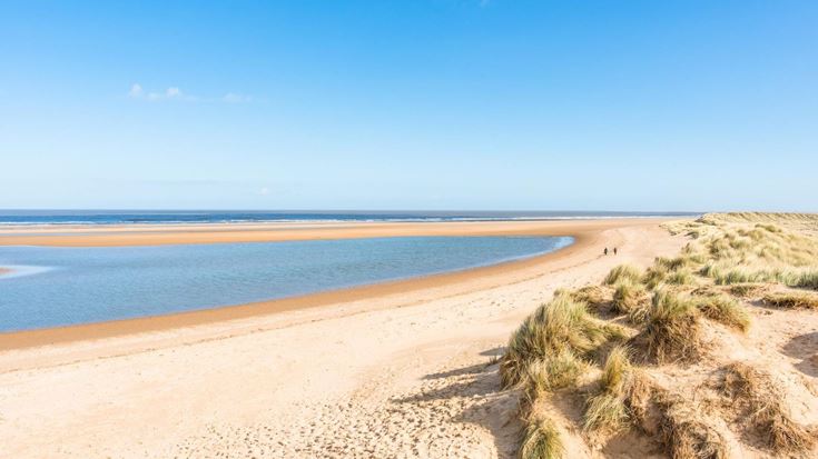 A long sandy beach along the Norfolk Coastal Path, with grass along the shoreline.