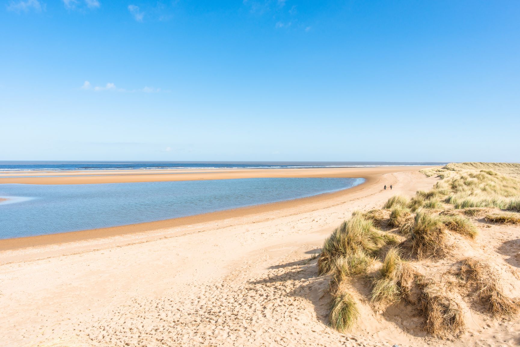 A long sandy beach along the Norfolk Coastal Path, with grass along the shoreline.