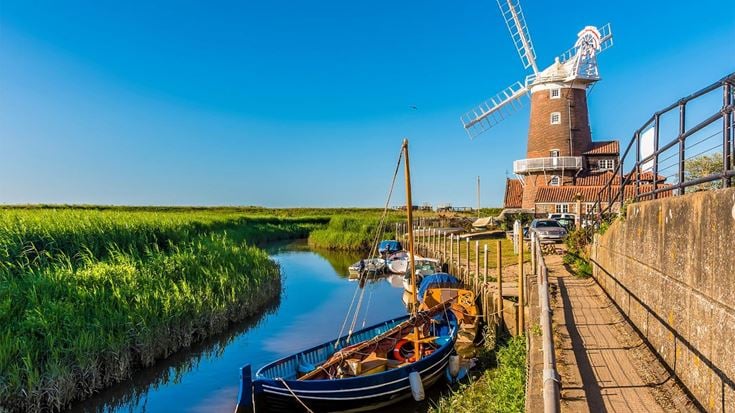 A small sailboat on the River Glaven in Cley, Norfolk, beside a windmill.
