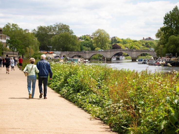 A senior couple walking on a path next to the River Thames with a bridge in the distance.