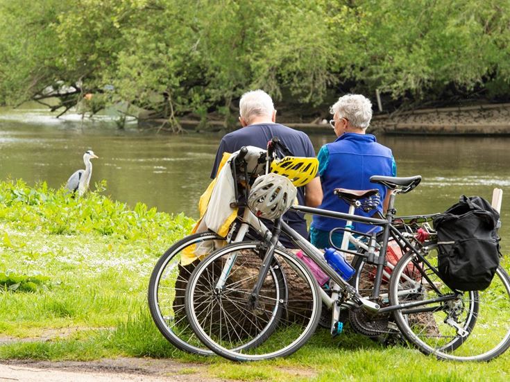 Two retired people rest on a log after riding their bicycles to a riverbank populated by birds.