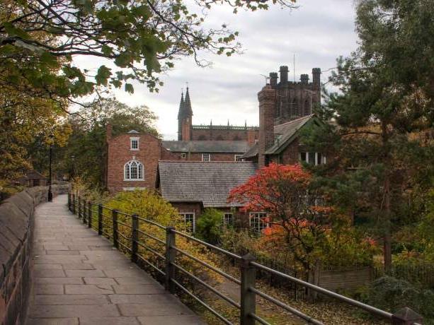 A path runs alongside the Chester City Walls with Chester Cathedral, autumnal trees, and other brick buildings in the background.