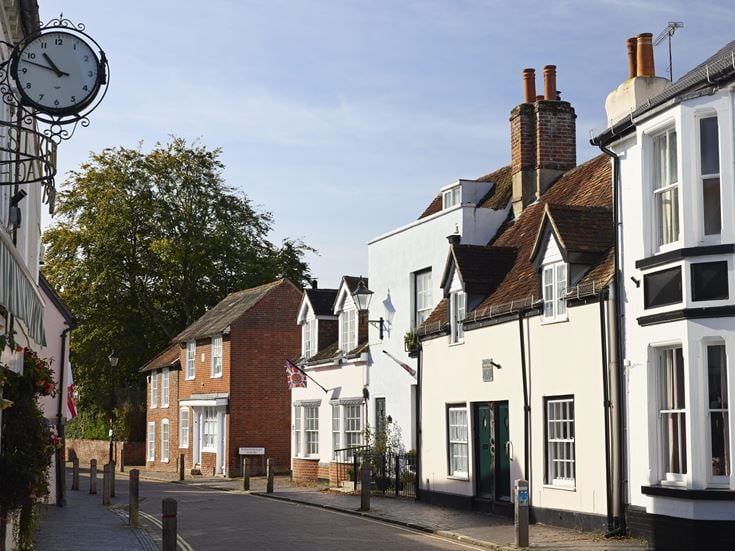 Traditional English houses, some with bay windows and white facades, and a clock line the road in a small Hampshire village.