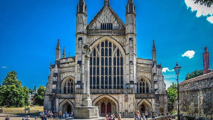 People walking or sitting outside the front exterior of Winchester Cathedral on a sunny day.
