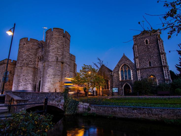 A bus at dusk passing by Westgate Towers in Canterbury, with the Great Stour river in the foreground.