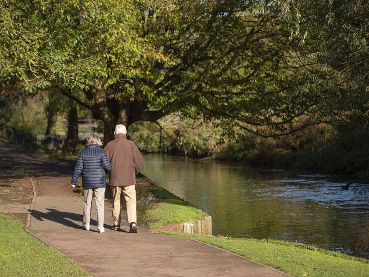 A man and woman walking on a path through Westgate Gardens next to the River Stour.
