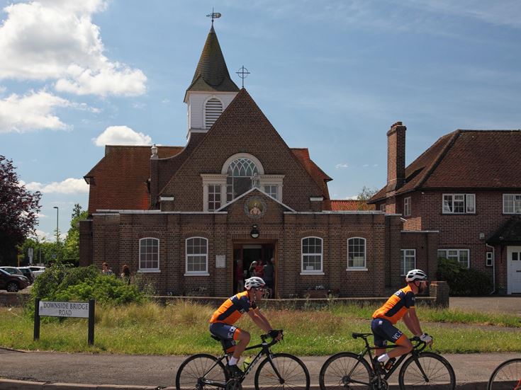 Two people on bikes cycling past a Sacred Heart Catholic Church on Downside Bridge Road, Cobham.