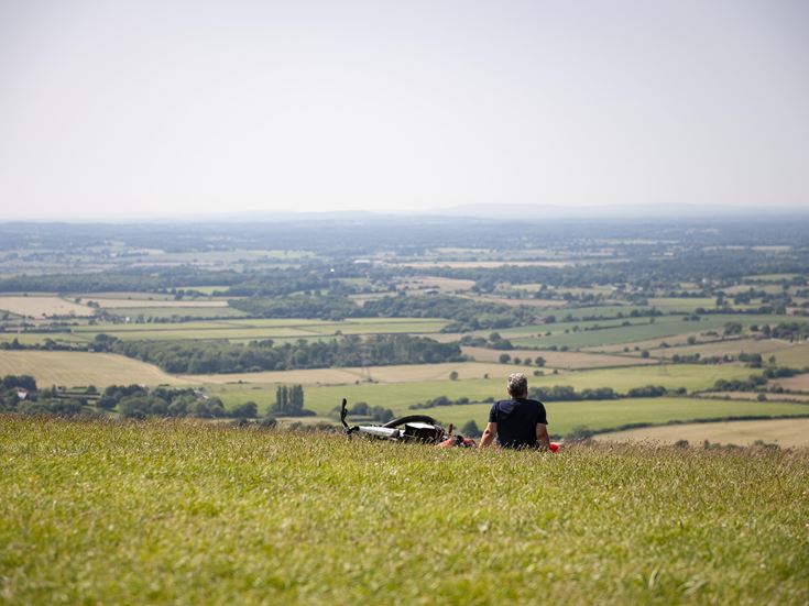 A person sits on a grassy hillside looking out over farming fields.