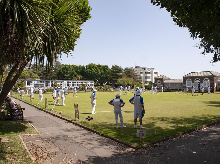 Several people stand on a boules pitch wearing traditional boules attire in the sun. The two closest to the camera are under the shade of a tree.