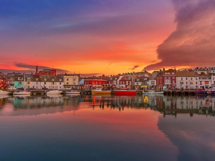 The coastal town of Weymouth in Dorset at sunset, with orange and purple clouds in the sky and buildings reflected in the water.