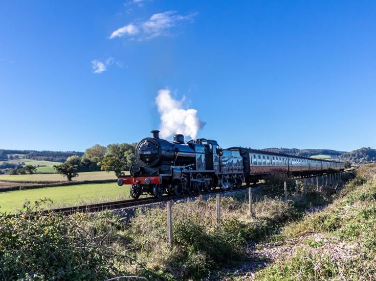 A vintage steam train and carriages on the West Somerset Railway with green fields and blue sky in the background.