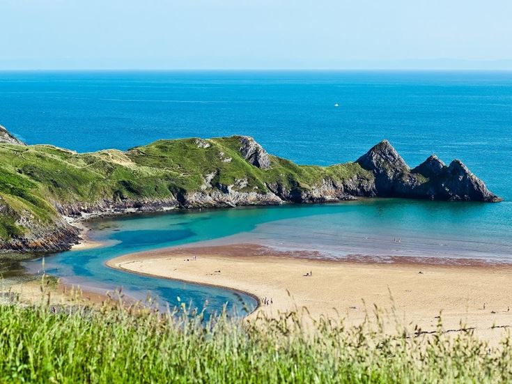 A sandy beach and clear waters surround Three Cliffs Bay in Wales.