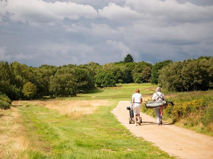 Two retirees carrying golf bags and clubs on a green.