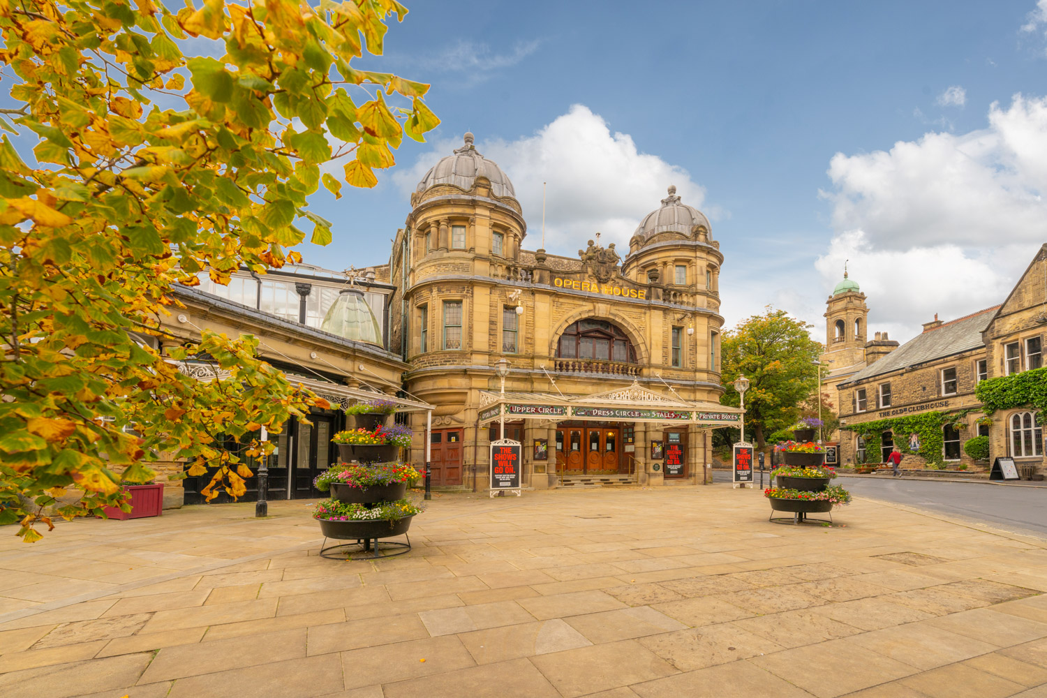 a large building with a dome and a courtyard with trees and flowers with Buxton Opera House in the background