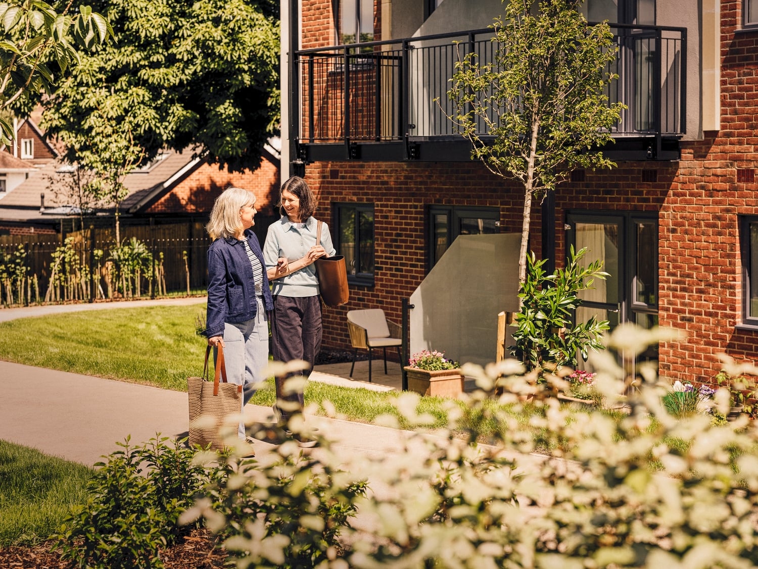 Two women walking on a path through gardens in front of a brick building.