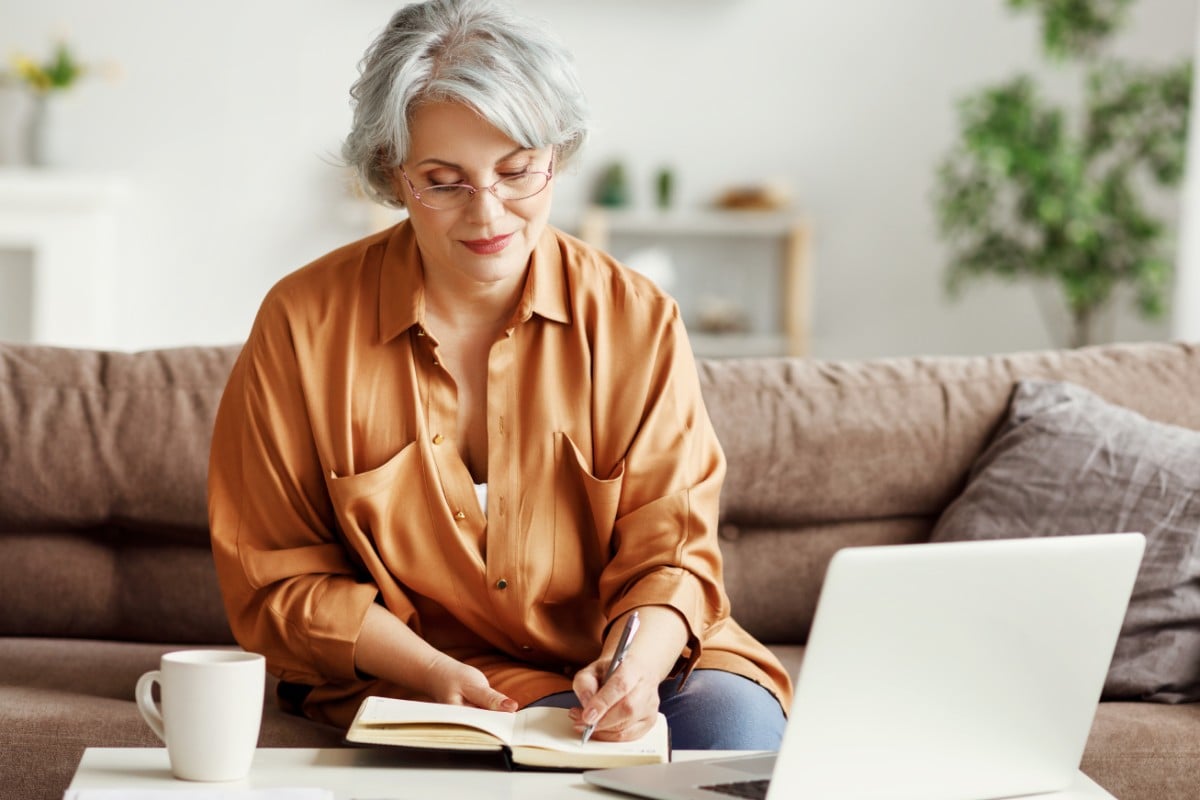 A retiree sitting on a sofa taking notes whilst working from their laptop