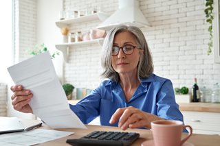 Senior woman reviewing finances with a calculator and paperwork at her laptop considering if pensioners pay tax