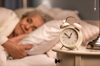 An older woman peacefully asleep in bed with an alarm clock in the foreground highlighting sleep needs for older adults.