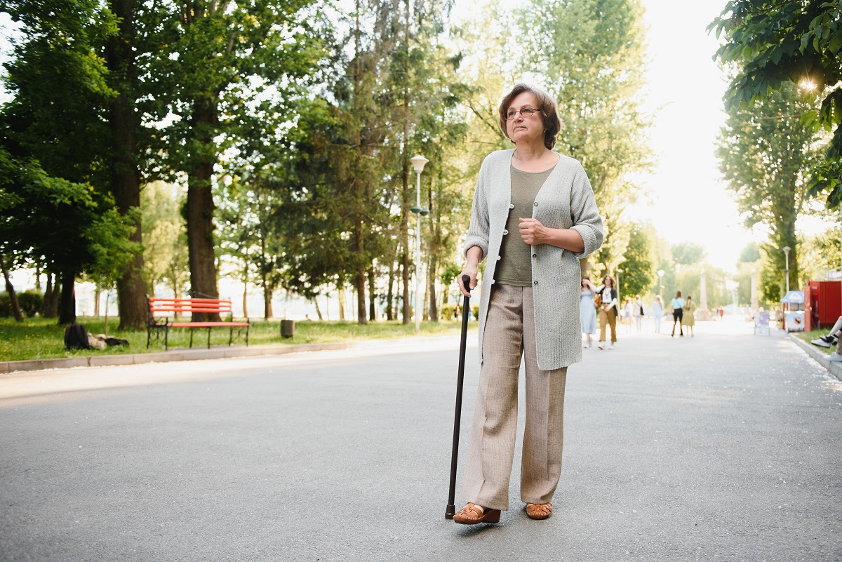 Over 60s retired woman walks in park with a walking stick.