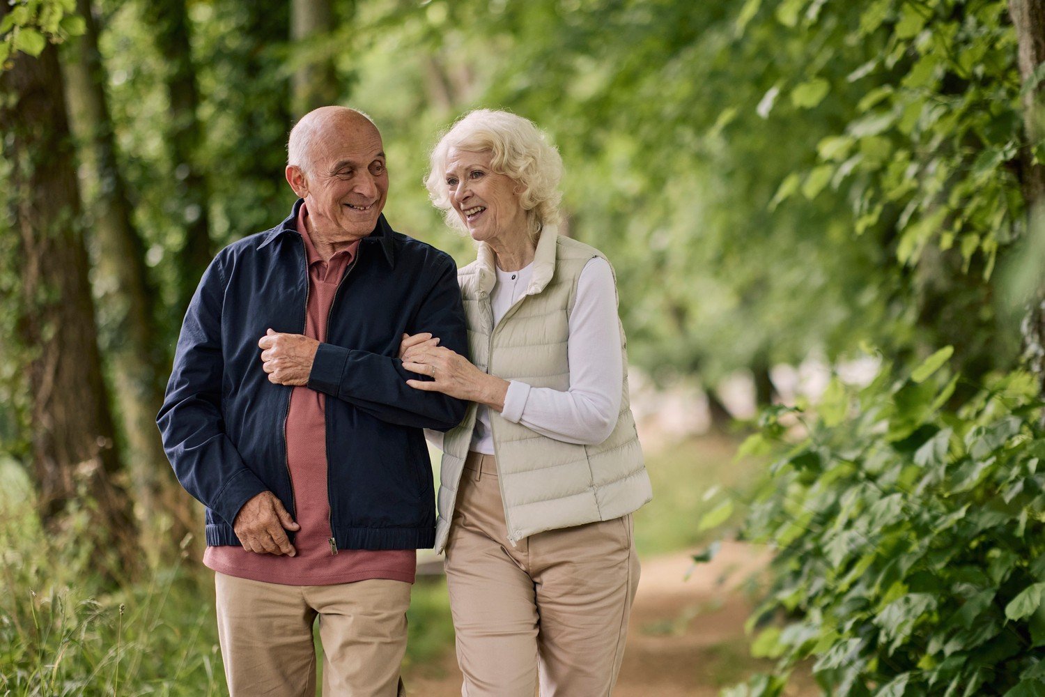 Man and woman linking arms, walking through green space and trees, near retirement apartments at a McCarthy Stone development