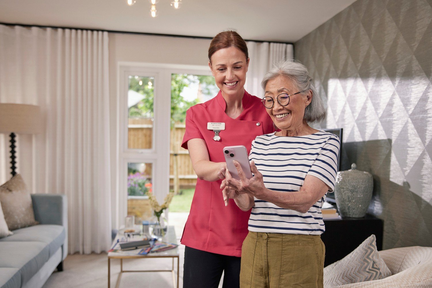 A retiree and McCarthy Stone team member, both smiling at the phone held in the retiree’s hand. They stand together in the retiree’s retirement home.