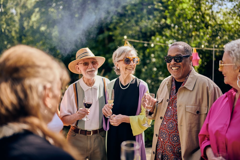 A group of older people enjoying a garden party, with various drinks in hand and smoke from a barbecue pluming overhead