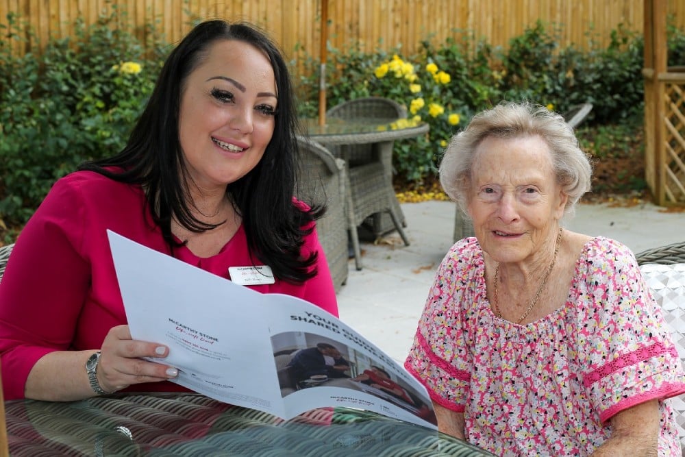 McCarthy Stone Sales Consultant Kelly sits with a retiree, Christine. The Sales Consultant holds a ‘Shared Ownership’ brochure to explain the concept of Older Persons Shared Ownership