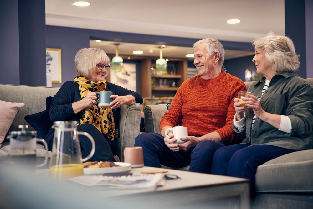 Three older people sat around a table, enjoying a catch-up in the communal lounge of a McCarthy Stone retirement development