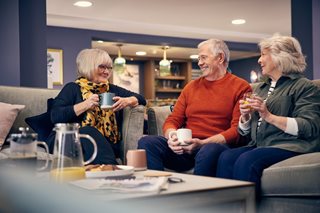 Three older people sat around a table, enjoying a catch-up in the communal lounge of a McCarthy Stone retirement development