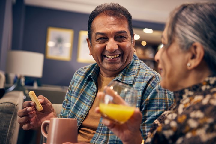 Two people smiling and engaging with each other whilst drinking beverages and eating biscuits.