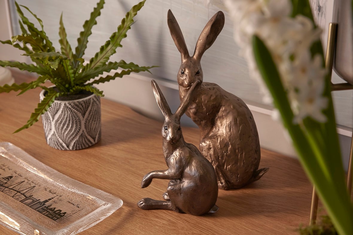 A close-up of two brass rabbit statues on a wooden table, surrounded by indoor plants.