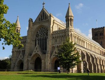 a large stone building with a fence in front of it with St Albans Cathedral in the background