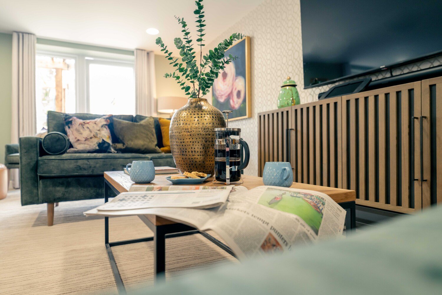 A cafetière, two mugs and a newspaper atop a coffee table in a retirement development lounge.