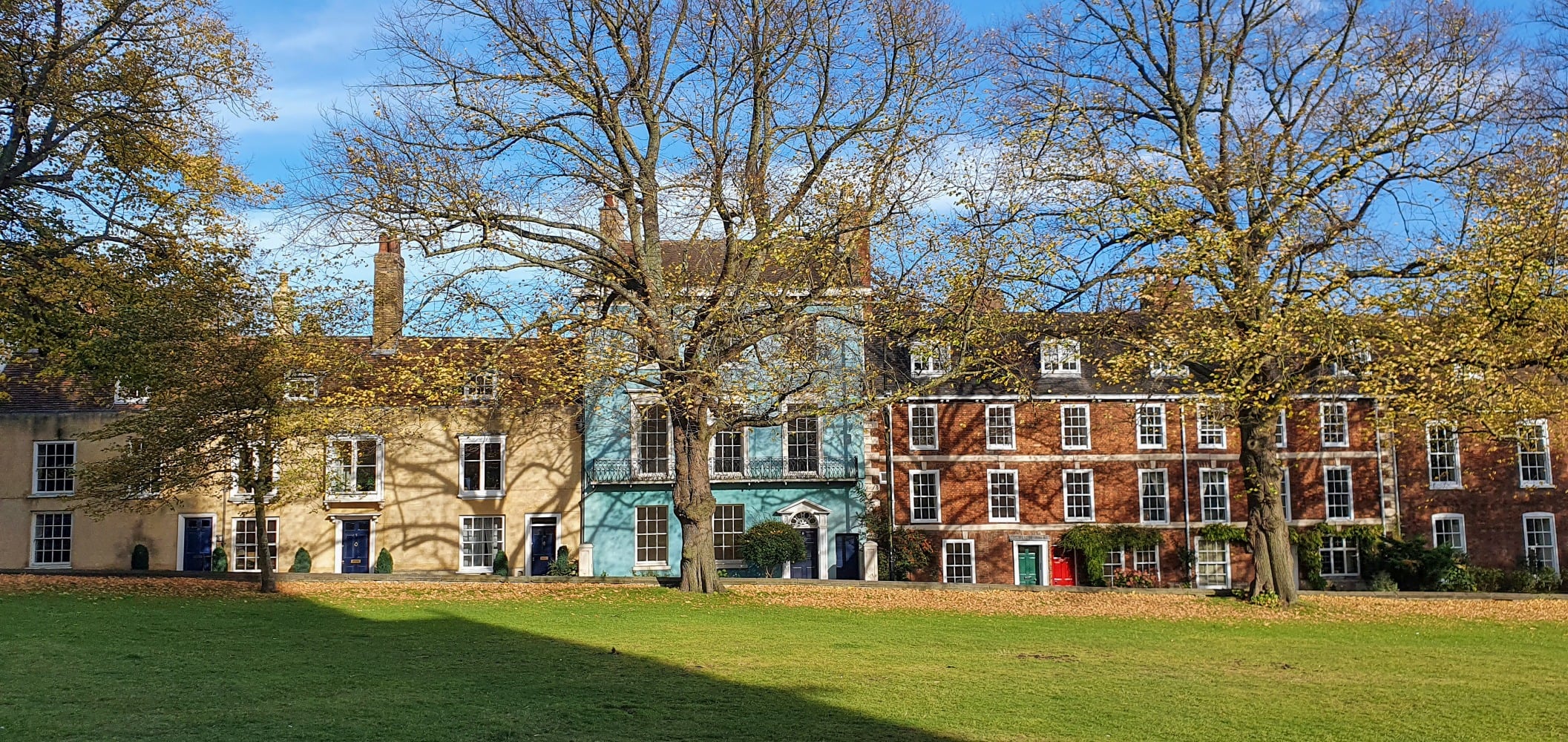 A row of historic buildings in Lincoln along a tree lined street, next to a green field.