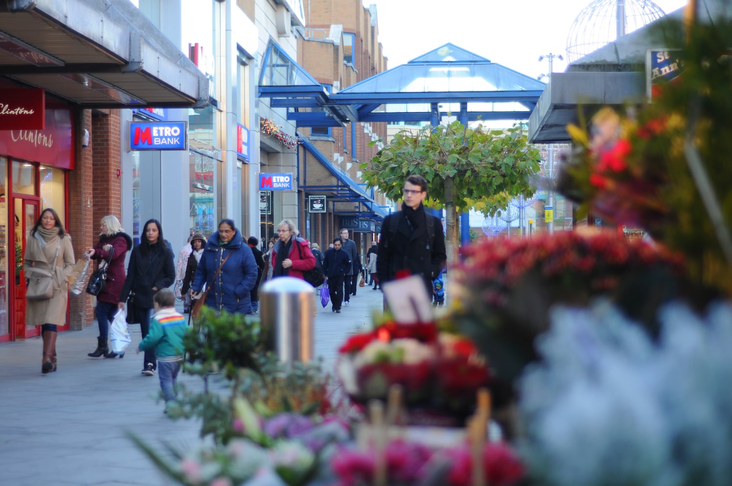 People walking past shops in Harrow town centre, with flowers in the foreground.