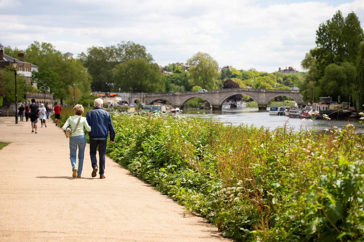 A senior couple walking on a path next to the River Thames with a bridge in the distance.