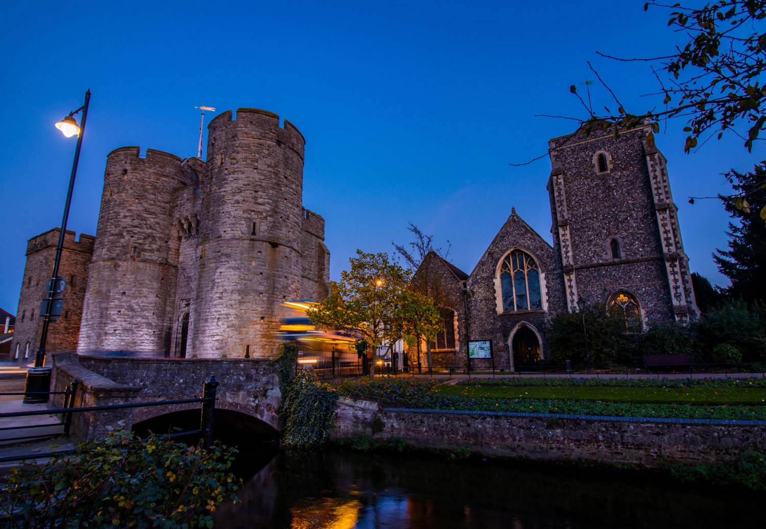 A bus at dusk passing by Westgate Towers in Canterbury, with the Great Stour river in the foreground.