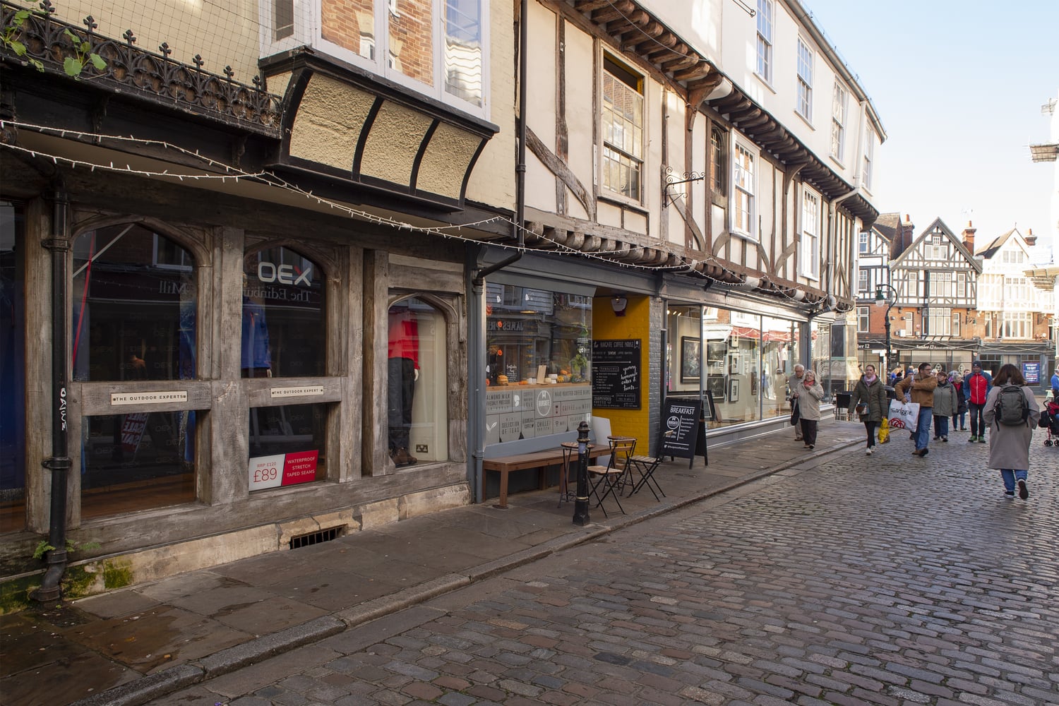 A group of people walking on a cobbled street next to a Tudor style building.
