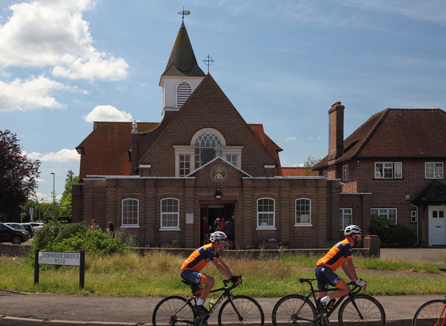Two people on bikes cycling past a Sacred Heart Catholic Church on Downside Bridge Road, Cobham.